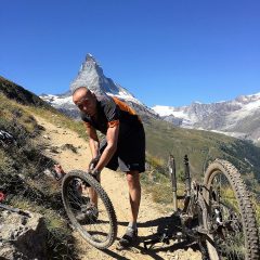 Mountain biker fixing a tyre on a rocky Alpine trail, a snowy peak under blue sky behind