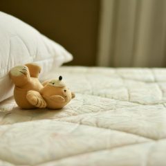 A teddy bear lying on a soft white quilted mattress topper beside a pillow