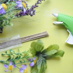 A green glue gun beside faux flowers and floristry wire on a yellow-green board