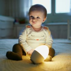 A baby sitting on a bed beside a glowing egg-shaped night light
