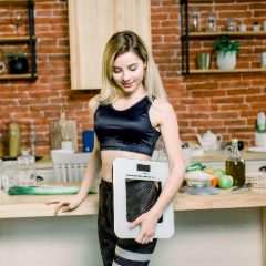 A smiling woman in sportswear holding a white digital scale in her kitchen