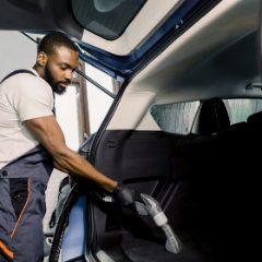 Smiling man in grey work overalls vacuuming the boot of a car with a hose and wide nozzle