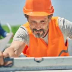 Smiling builder in an orange hard hat and hi-vis vest laying a long spirit level on a block wall outdoors