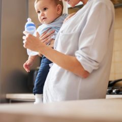 Smiling mother holding her baby, who grips a milk bottle, in a bright kitchen