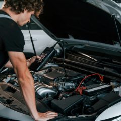 Young man leaning into an open engine bay holding a small black handheld unit wired to the battery