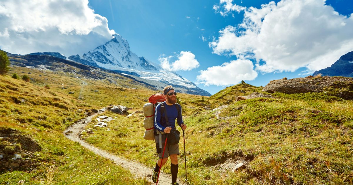 A hiker with trekking poles on a grassy Alpine path below a snowy peak