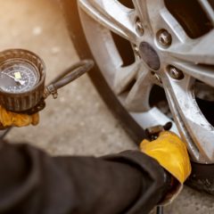 Gloved hand holding a round dial tyre gauge in a rubber guard on a short hose while the chuck sits on a car's valve