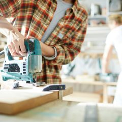Teal and black jigsaw cutting a board on a workbench, a person in a red checked shirt holding it