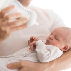 Father in a white T-shirt cradling his sleeping newborn and holding a clear baby bottle, in soft bright light