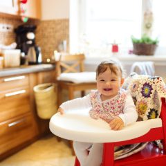 Smiling baby girl in a floral top sitting at the round white tray of a high chair in a sunny kitchen