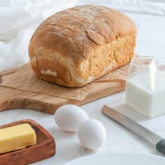 Home-baked white loaf on a wooden board with a carton of milk, eggs, butter and a bread knife on white marble