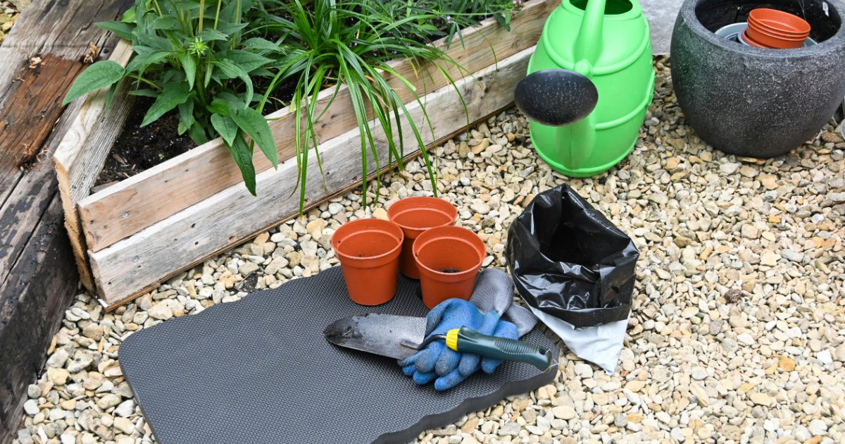 A gray kneeling mat on gravel beside pots, gloves and a watering can