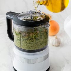 White compact food processor with a clear bowl of green pesto, olive oil being poured in through the lid