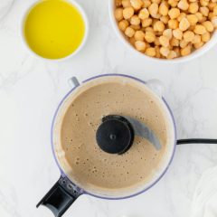 Food processor bowl of smooth hummus seen from above, beside bowls of olive oil and chickpeas on white marble