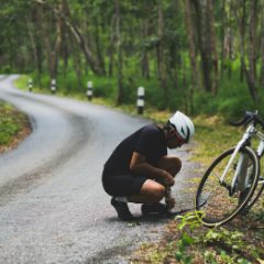 Cyclist in a white helmet crouched fixing a road bike at the edge of a winding forest road