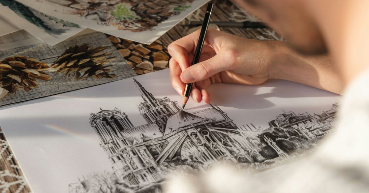 A hand drawing a detailed cathedral in pencil, with sketches and pine cones on the table behind