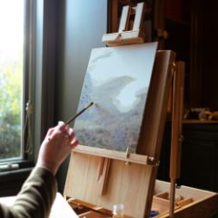 Hand painting a landscape on a canvas held by a wooden box table easel beside a window