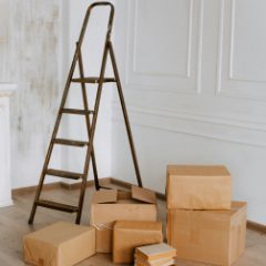 Dark metal step ladder with a top hand loop standing open in a bright white-panelled room beside moving boxes and a stack of books