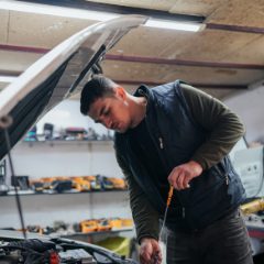 Young mechanic in a navy gilet leaning under an open car bonnet in a bright garage, checking the engine