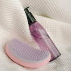 Pale pink palm detangling brush with lilac teeth lying on a cream knitted throw beside a pink pump bottle