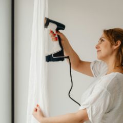A woman steaming a white curtain with a black handheld steamer