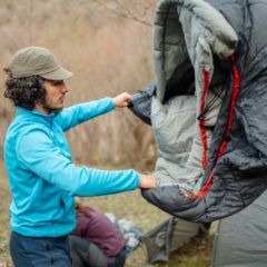 A camper holding up a grey mummy sleeping bag with a red zip