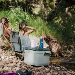 Children in camping chairs by a river beside a sage green cool box