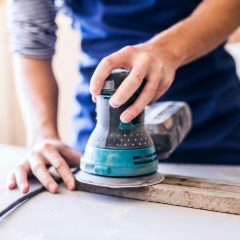 Hand on a teal and black electric sander smoothing a wooden board on a white workbench