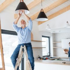 Smiling older man on a step ladder fitting a black pendant light in a bright new kitchen