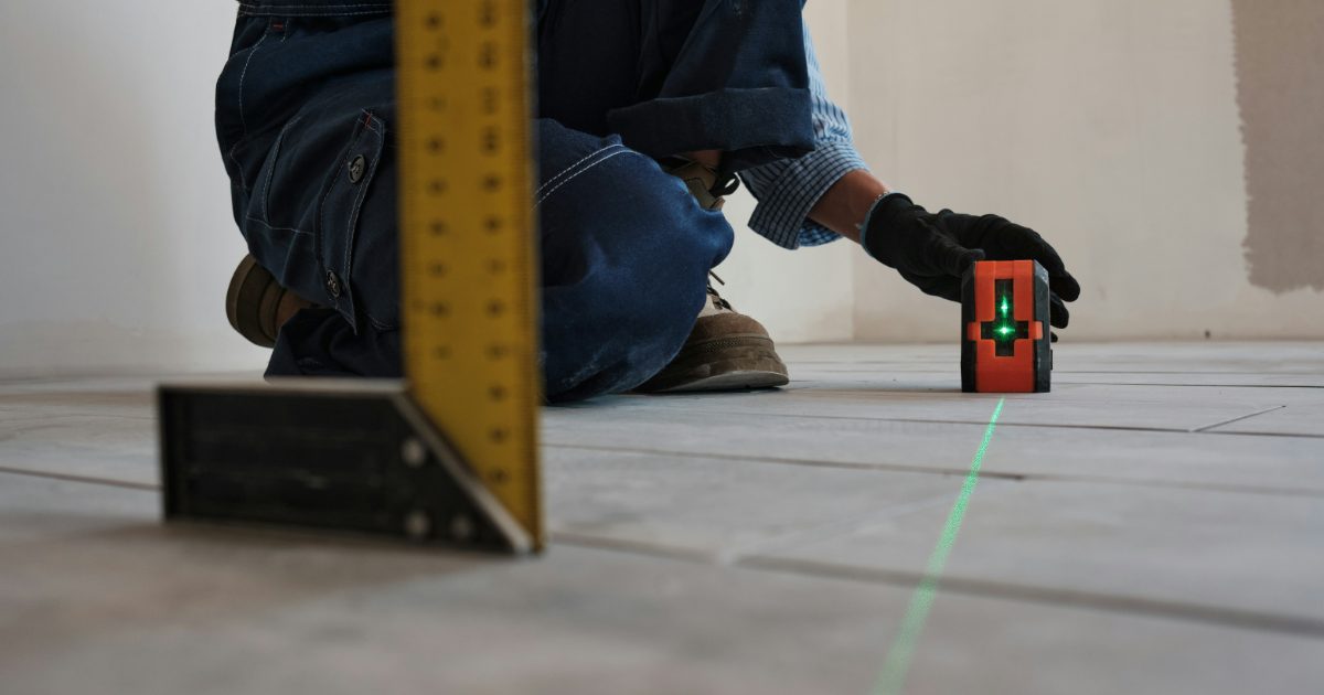A tiler kneeling on a new floor beside a small cross-line laser projecting a green line