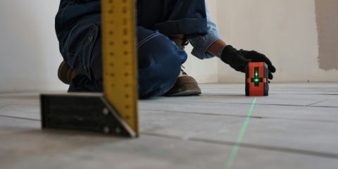 A tiler kneeling on a new floor beside a small cross-line laser projecting a green line