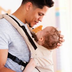 Father in a striped T-shirt smiling at his toddler son facing him in a cream front carrier with black buckles