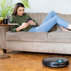 A woman relaxing on the sofa with her phone while a black robot vacuum cleans