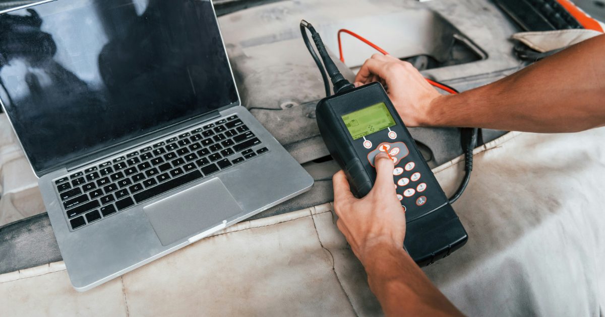 Hands using a handheld car diagnostic scanner beside a laptop