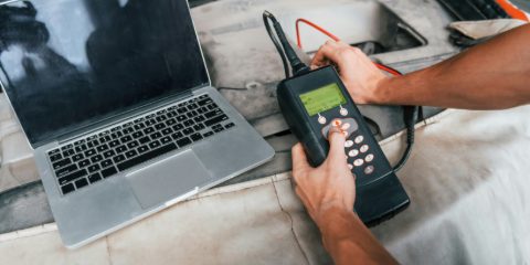 Hands using a handheld car diagnostic scanner beside a laptop