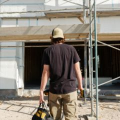 Builder in a cap walking towards scaffolding carrying a black and yellow tool bag