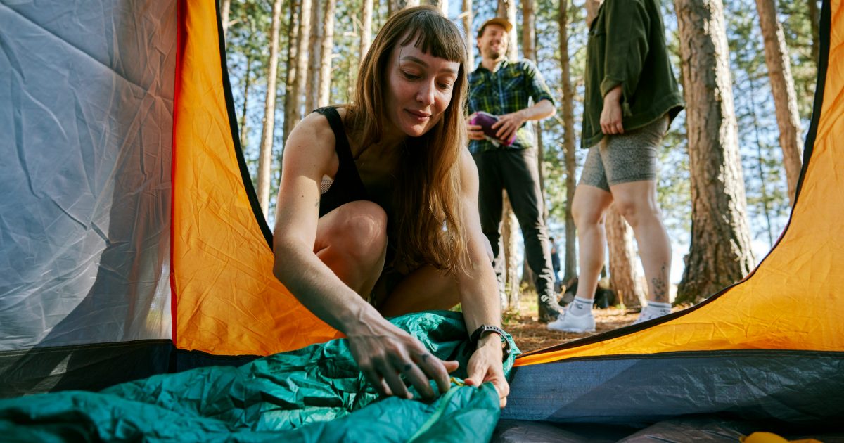 A woman laying out a green sleeping bag in a tent in a pine wood, friends behind her