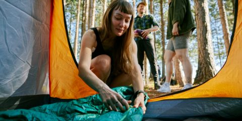 A woman laying out a green sleeping bag in a tent in a pine wood, friends behind her