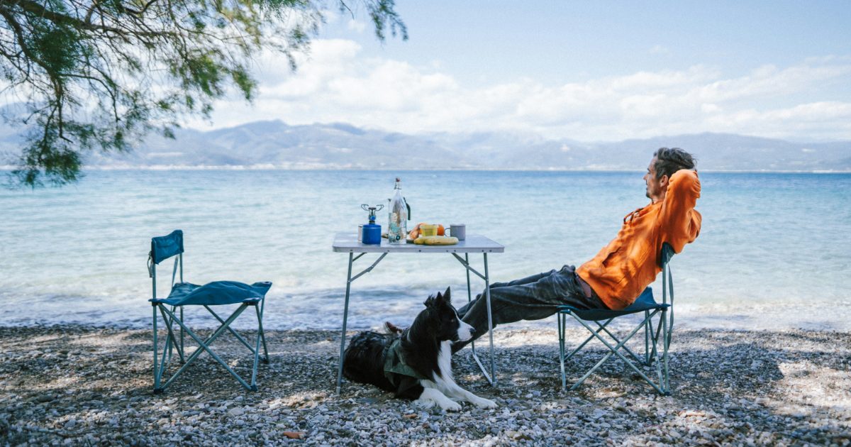 A man relaxing in a camping chair by a lake, his dog at his feet and a camping table beside him