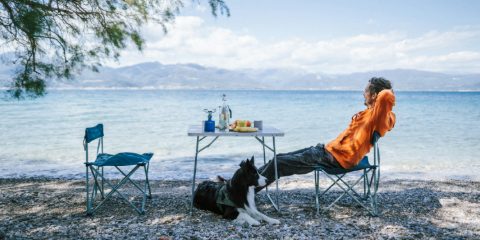 A man relaxing in a camping chair by a lake, his dog at his feet and a camping table beside him