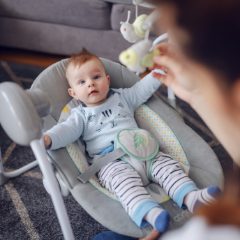 Baby in a striped outfit in a grey rocker seat with a toy mobile, his mother playing with him on a rug