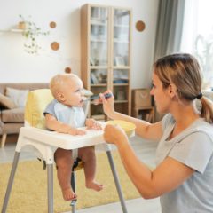 Mother kneeling to spoon-feed her smiling baby son in a white high chair in a bright living room