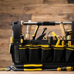 Black tool tote with yellow trim and rows of pockets full of hand tools on a wooden bench