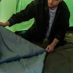 A man zipping up a grey sleeping bag inside a green tent