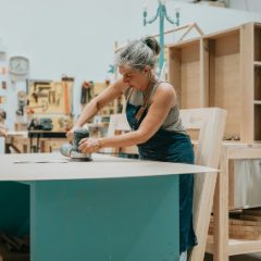 Smiling grey-haired woman sanding a table top with a random orbit sander in a bright workshop