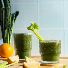 Two ribbed glasses of fresh green juice with celery, cavolo nero, an orange and lemon halves on a wooden board