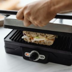 Hand lowering the stainless steel lid of a black sandwich grill onto a sandwich, on a white worktop