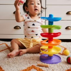 A toddler sitting on a rug beside a rainbow ball run tower