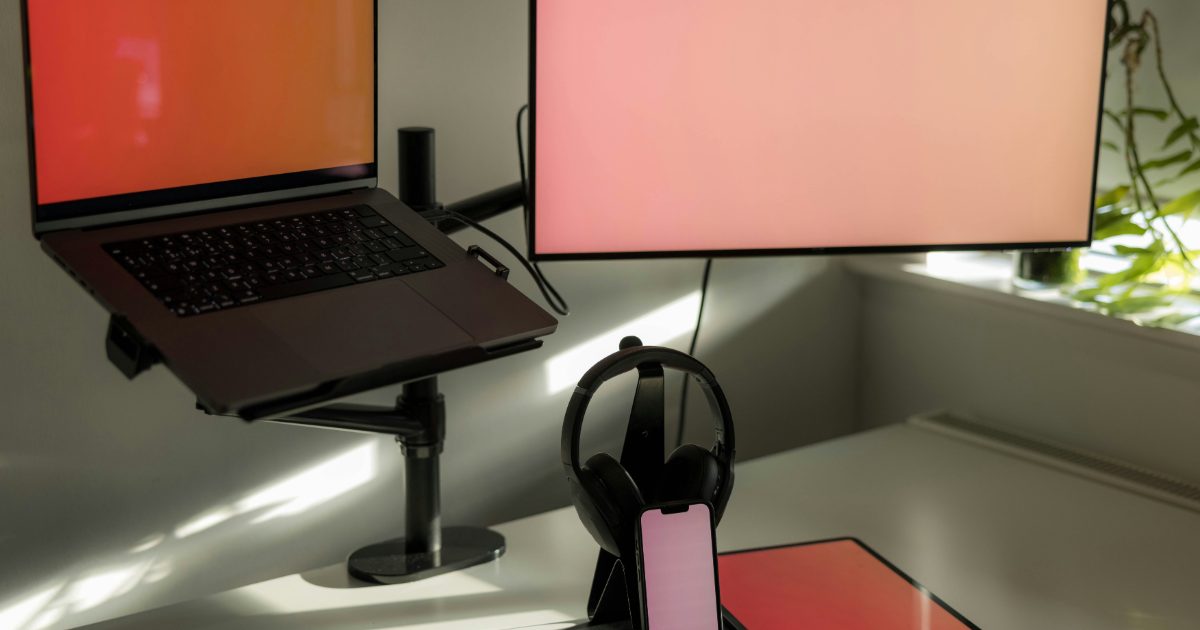 A monitor and a laptop tray held above a bright white desk on a black pole-mounted arm, with headphones on a stand and a phone below
