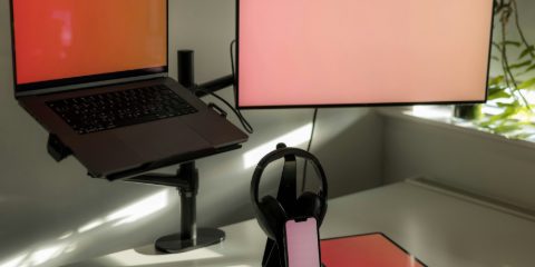 A monitor and a laptop tray held above a bright white desk on a black pole-mounted arm, with headphones on a stand and a phone below